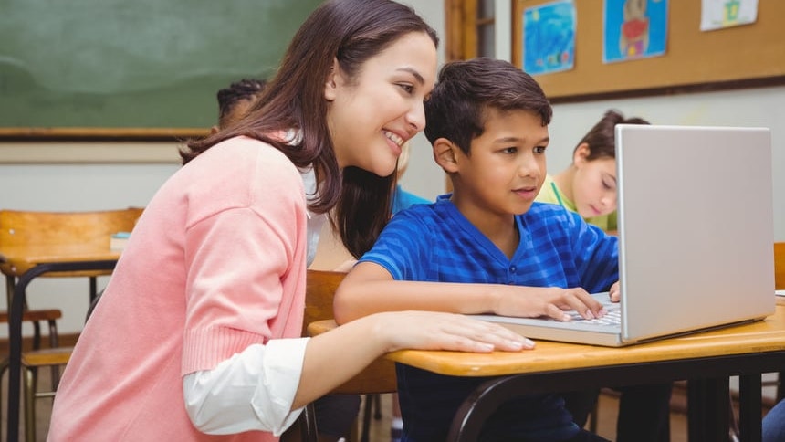 A female teacher looking over the shoulder of a young male (assumed) student looking at a laptop.